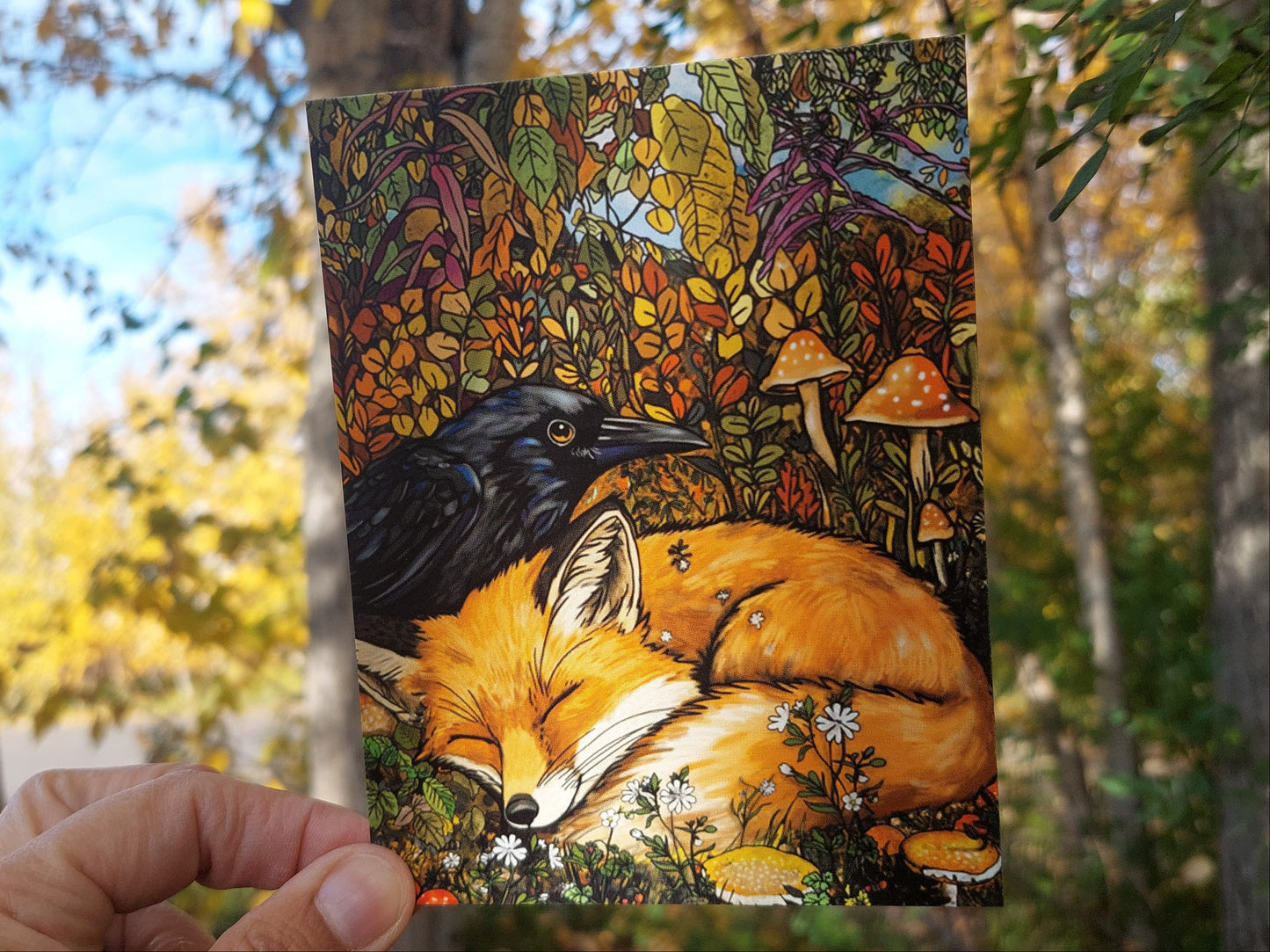 Hand holding a colorful illustration of a fox and black bear against a natural background with trees and leaves.