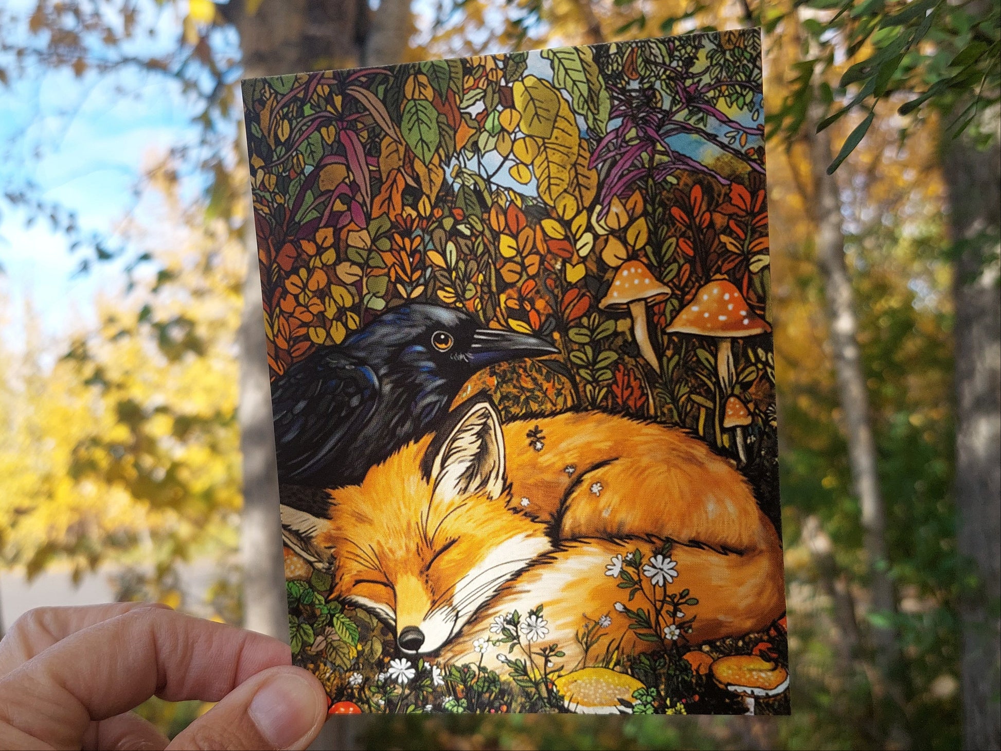 Hand holding a colorful illustration of a fox and black bear against a natural background with trees and leaves.